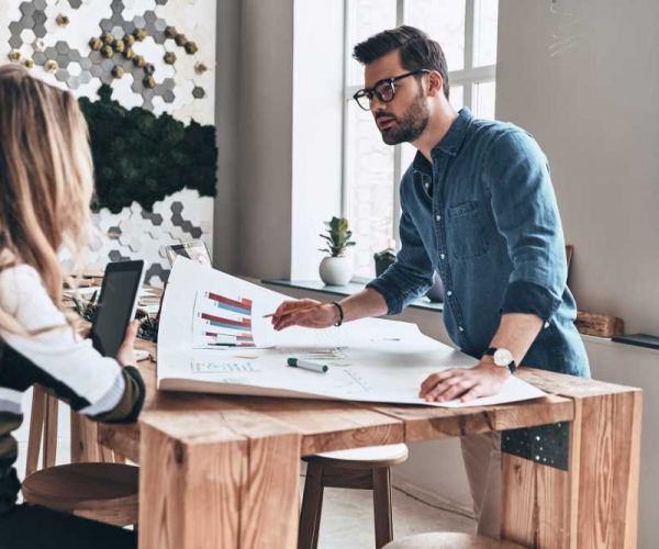 Discussing strategy. Two young modern business people in smart casual wear using blueprint while working in the creative working space Discussing strategy. Two young modern business people in smart casual wear using blueprint while working in the creative working space